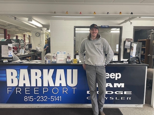 Photo is of a man wearing a grey sweatshirt and pants and a baseball cap. He is standing in front of acounting with a sign that says Barkau Freeport. The photo introduces a job training success story.