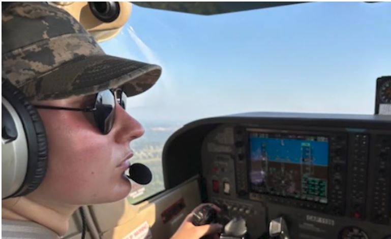 Photo is the profile of a young woman in a camoflage baseball cap style hat flying a plane. Story introduces a Ruthie's success story.