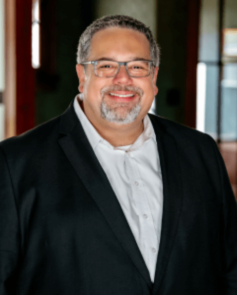 Photo is of a smiling man with greying hair, mustache and trimmed beard, black dress jacket and white dress shirt.