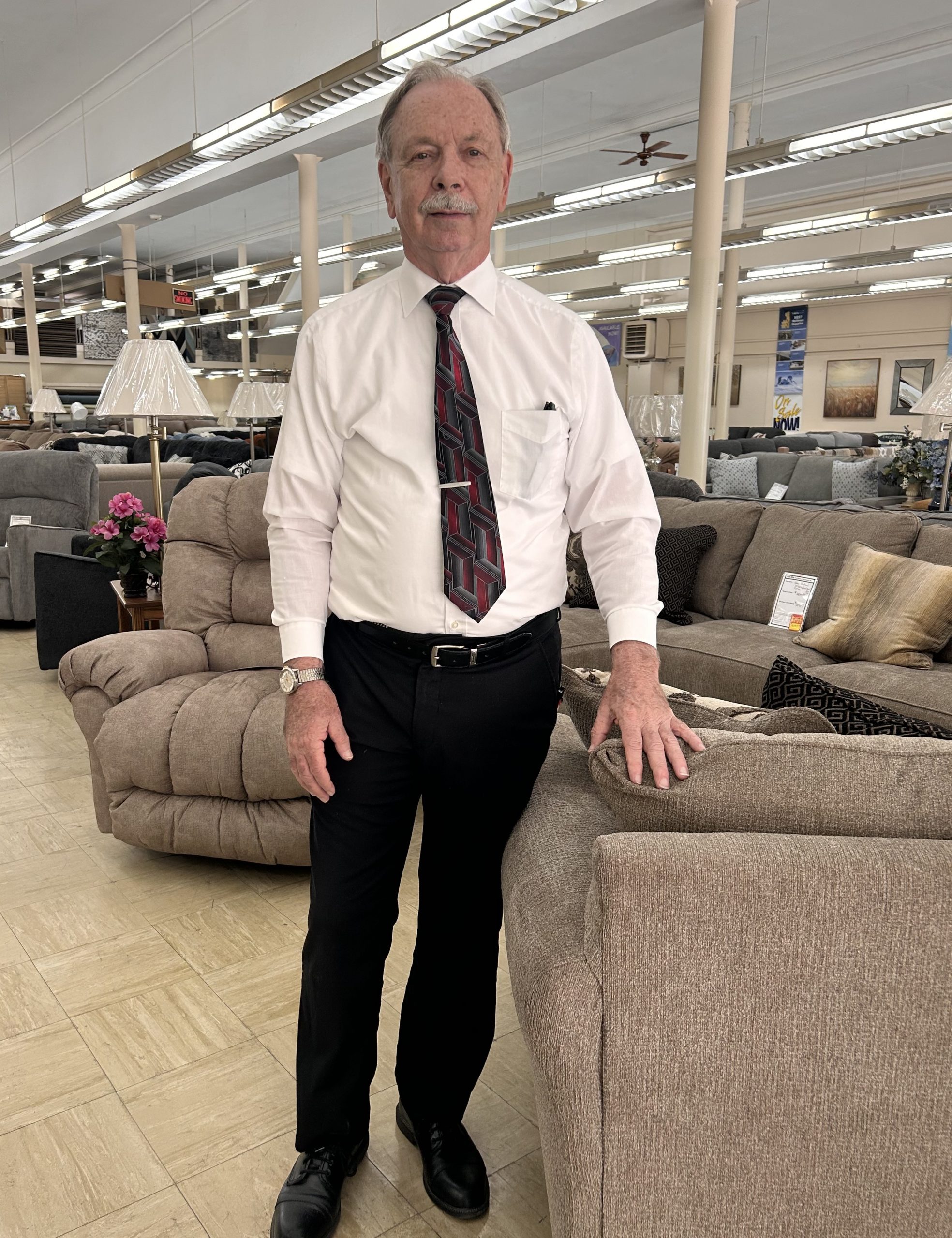 Photo introduces a part of the story about Rite-Way and features a man who is standing and wearing dark dress pants, a dress shirt, and tie. The man is standing in front of furniture displays including couches and chairs.