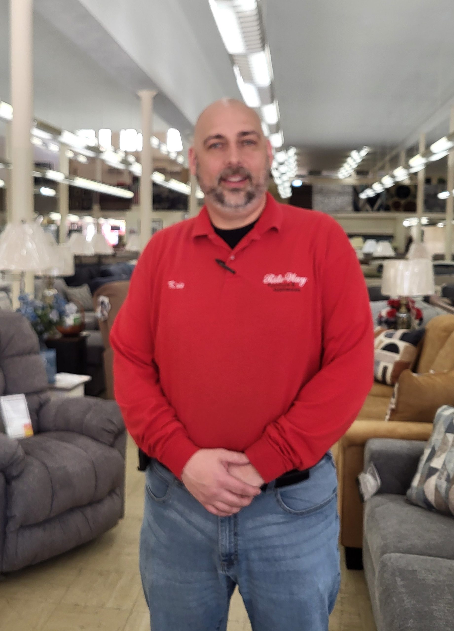 Photo of job training, Kristopher, wearing a red shirt and jeans and standing in a furniture store.