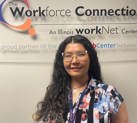 Smiling young woman earing a patterned colorful shirt, glasses and is standing in front of a clear plexiglass sign that has The Workforce Connection logo on it.