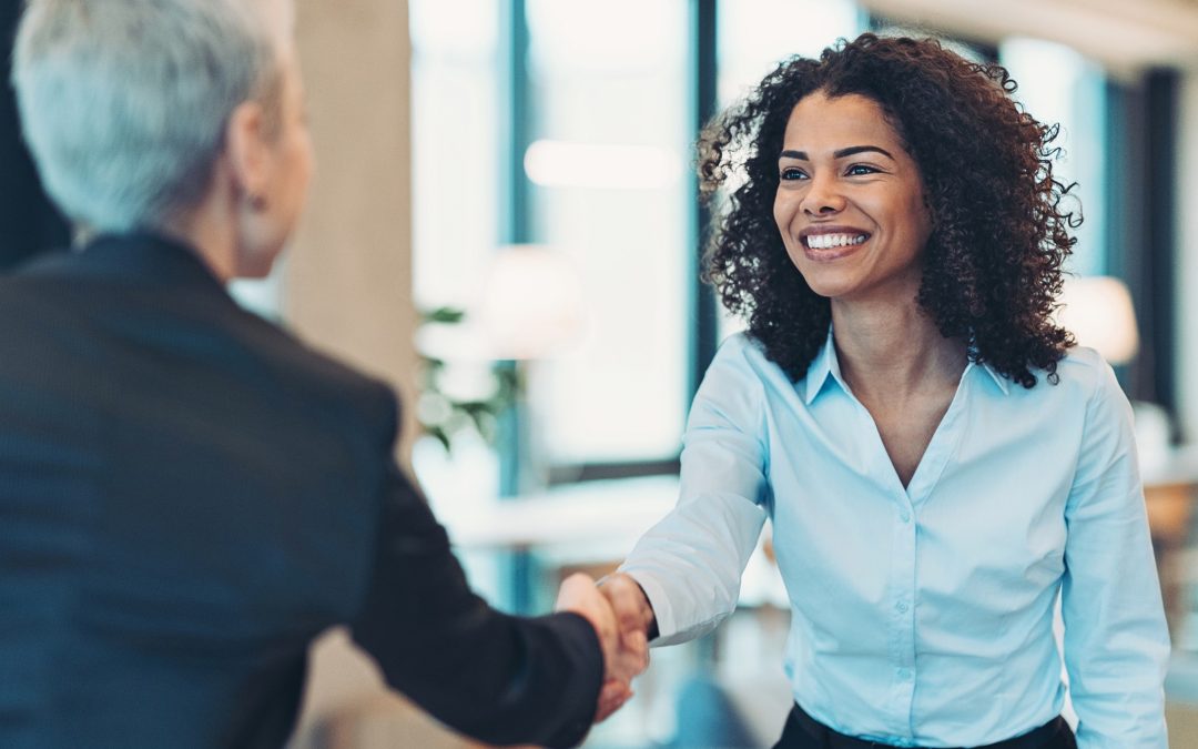 Photo shows a smiling woman with dark brown curly hair shaking the hand of another person in a business suit.